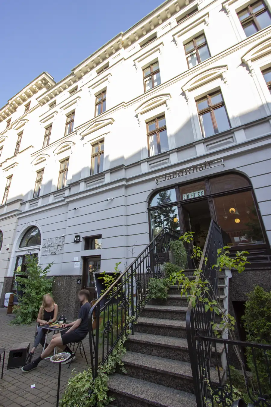 Summer facade with people dining at outdoor table, Landschaft entrance with iron staircase, full building height visible, blue sky