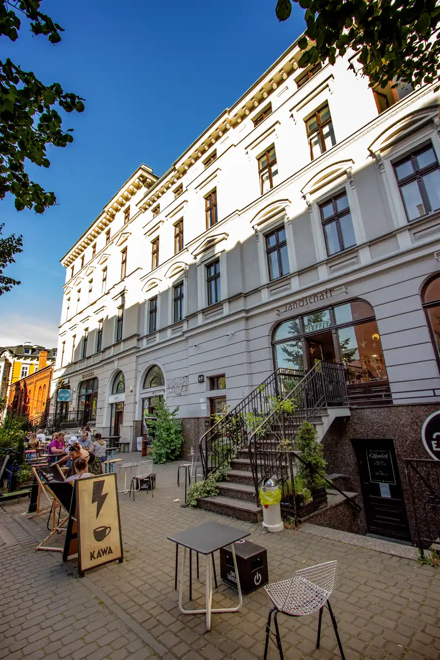 Street view with KAWA coffee sign, chair on pavement, full building height, Landschaft signage, blue sky, angled perspective