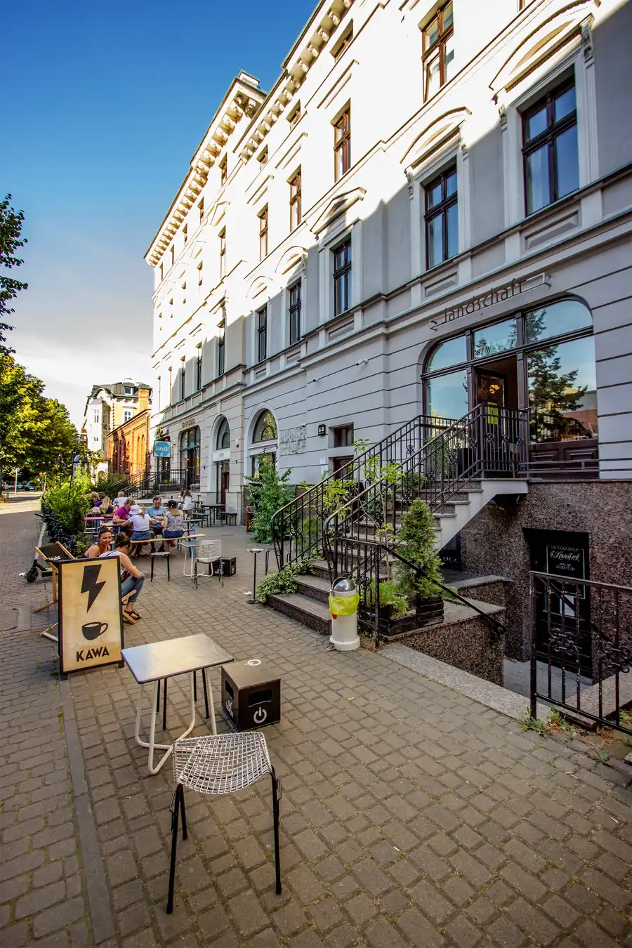 Building facade with KAWA sign, cafe entrance, wrought iron stairs, full four-storey height, trees, blue sky