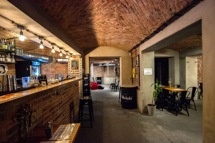 Basement bar interior with exposed brick vault ceiling, bar counter, Fritz-kola barrel table, industrial lighting, Tolix chairs