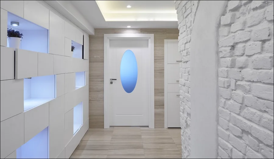 Spa corridor with white panelled wall, LED niches, and oval-glass door beneath brick ceiling