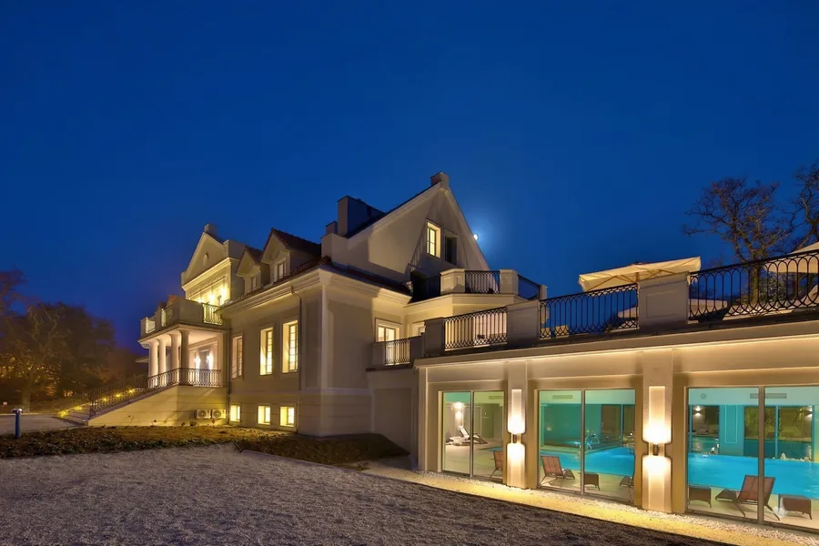 Night exterior revealing heritage manor architecture above and contemporary glass spa extension glowing below with pool visible