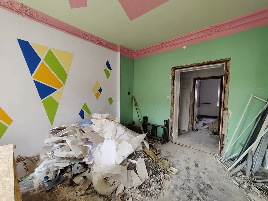 Before: pre-renovation room showing bare plaster walls, original timber window with deep reveal, radiator, and partially stripped floor surface