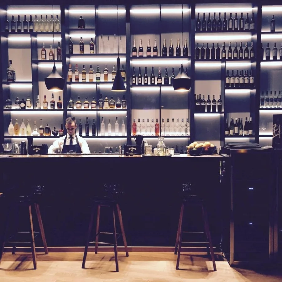 Interior: cocktail bar with bartender behind backlit bottle shelves, bar stools, and Tom Dixon pendants