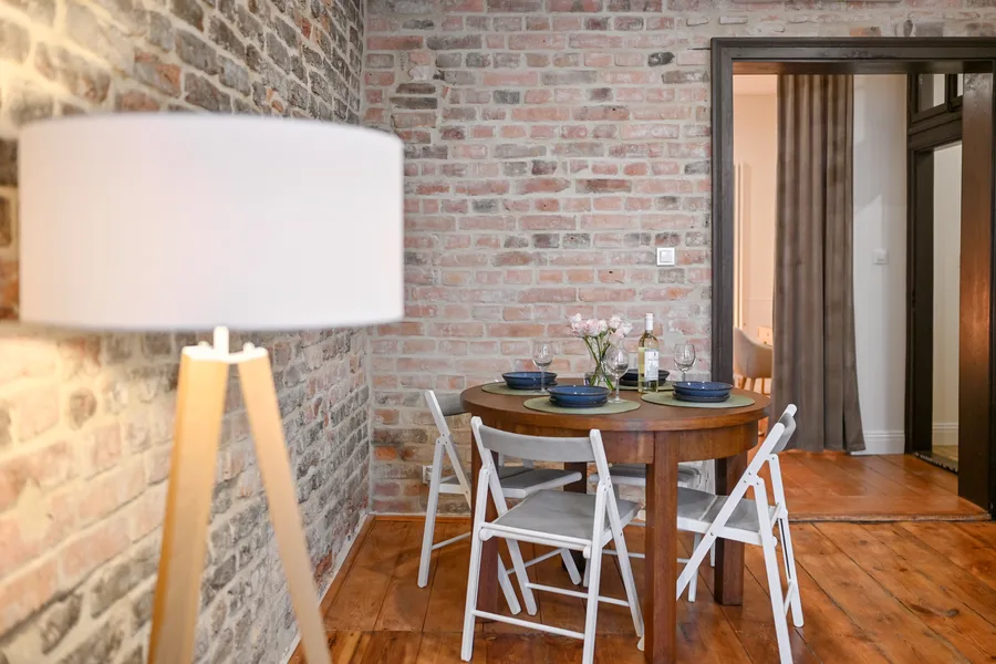 Interior: Apartment B kitchen-dining wide shot with exposed brick wall, timber open shelving, pendant lamp, and oak dining table with chairs