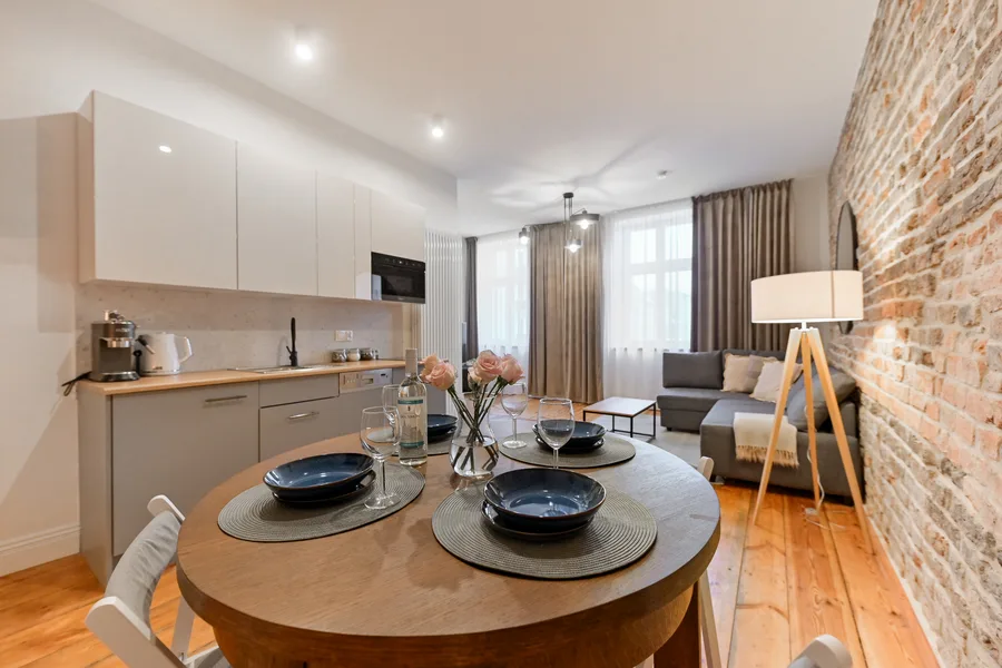 Interior: Apartment B dining area with exposed brick wall, warm pendant lamp illuminating oak table, and view toward kitchen behind