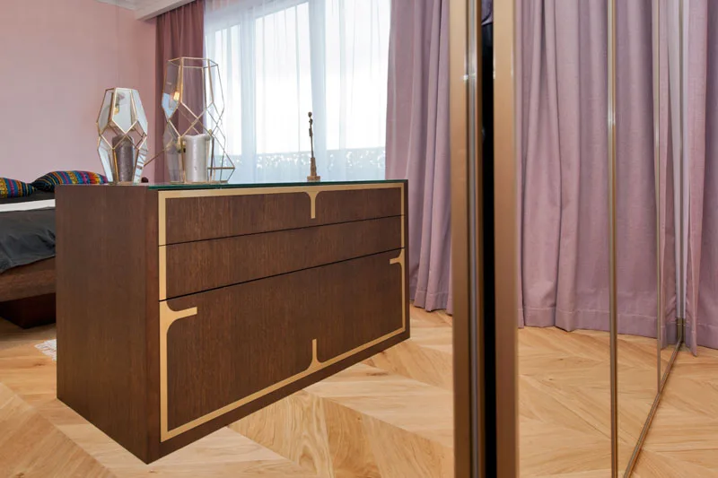Bedroom dresser detail showing custom dark wood with brass inlay patterns, geometric mirrored lamp, pink curtains, and mirror wardrobe edge