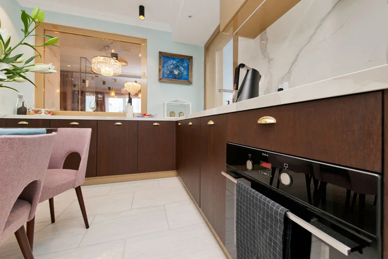 Kitchen counter detail with dark wood cabinets, brass cup handles, marble backsplash, built-in oven, and pink chair edge visible through glass partition
