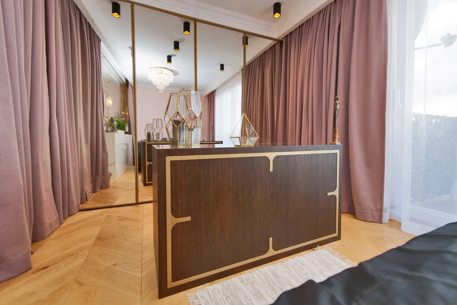 Bedroom with custom dark wood dresser with brass inlay geometric patterns, pink and mauve curtains, mirrored wardrobe doors, and herringbone parquet floor