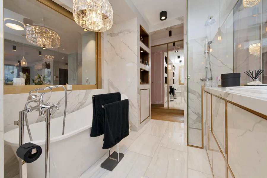 Bathroom alternate angle with freestanding tub from entry end, crystal chandelier, marble walls, wood-backed recessed niches, and glass shower partition