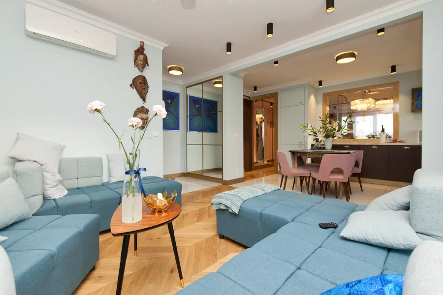 Living room panoramic with teal sofa, mid-century table, glass partition to kitchen-dining beyond, carved masks on wall, and pink chairs in distance