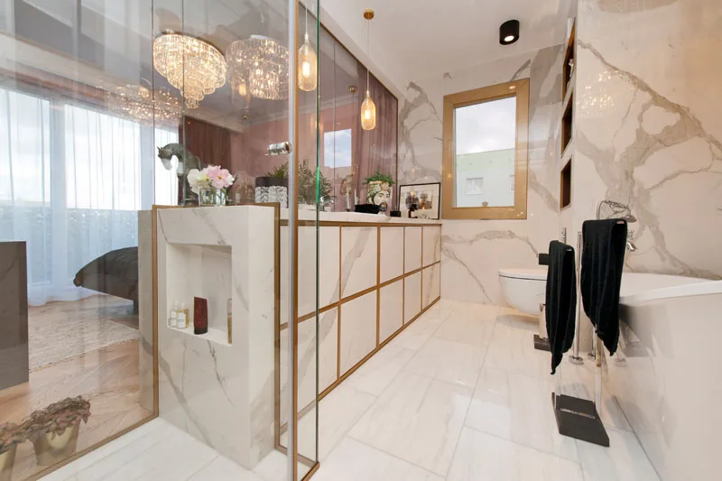 Bathroom viewed through glass partition showing marble walls, crystal chandelier reflection, and brass glass framing