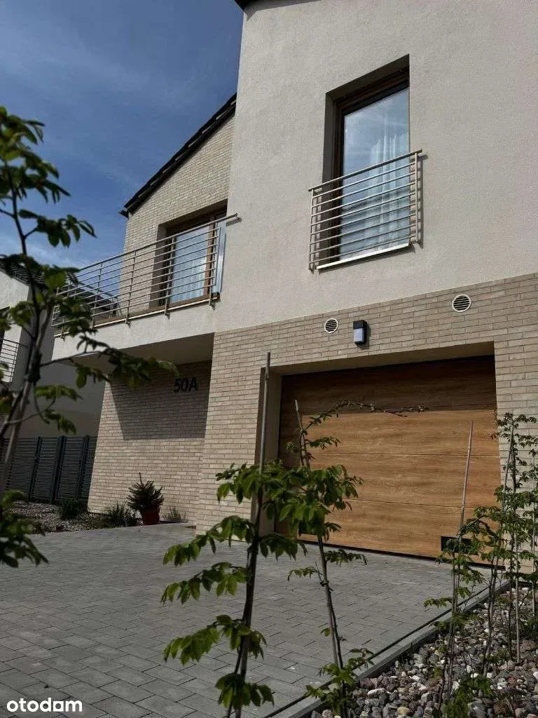 Close-up realization photo of entrance and garage showing wood-tone garage door, clinker brick wall, circular recuperation vents, and bronze window frames