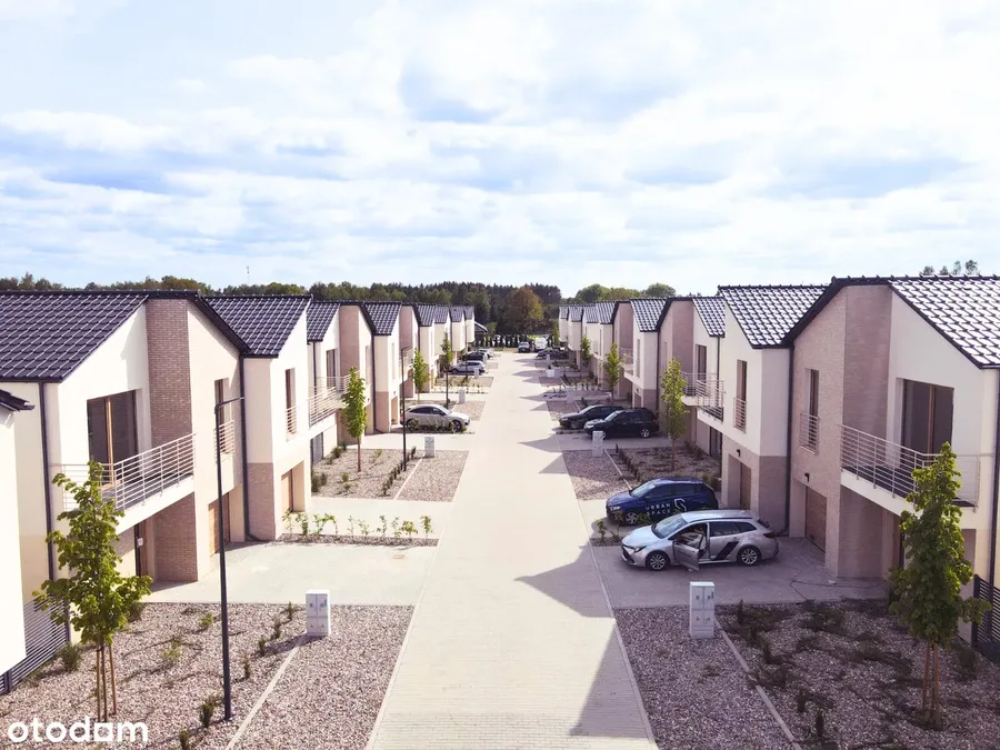 Drone photo along internal street axis showing two rows of semi-detached houses with gravel gardens, young trees, and parked cars in the gated community