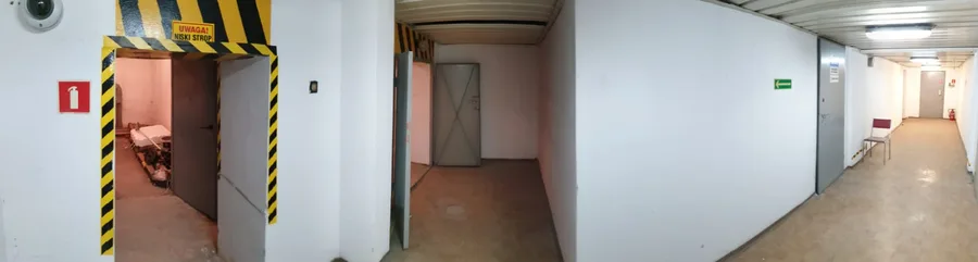 Documentary: corridor photograph with yellow-and-black hazard stripe warning sign reading Uwaga Niski Strop showing the low ceiling and raw industrial character of the existing 1975 building interior