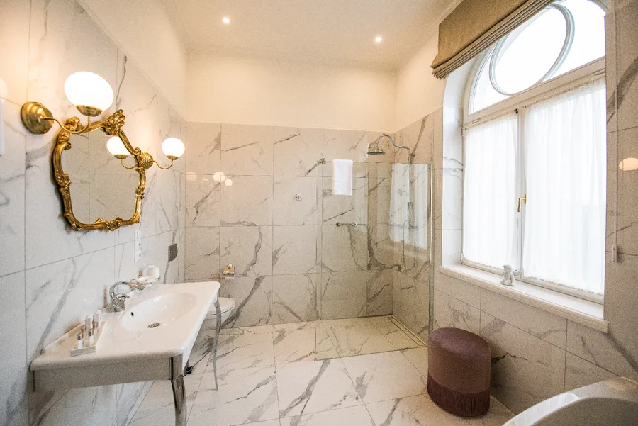Bathroom with marble walls, gold ornate mirror, walk-in shower, and arched window