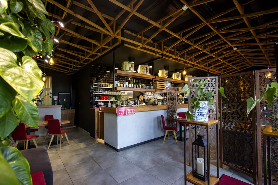Interior: bar counter overview with plants top-left, sake barrel shelf, metal screens, lattice ceiling