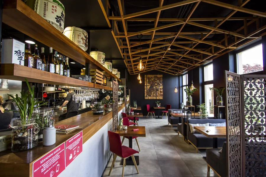 Interior: bar and sushi counter from dining floor — shelves with sake barrels, chef visible, Klimt, striped sofa