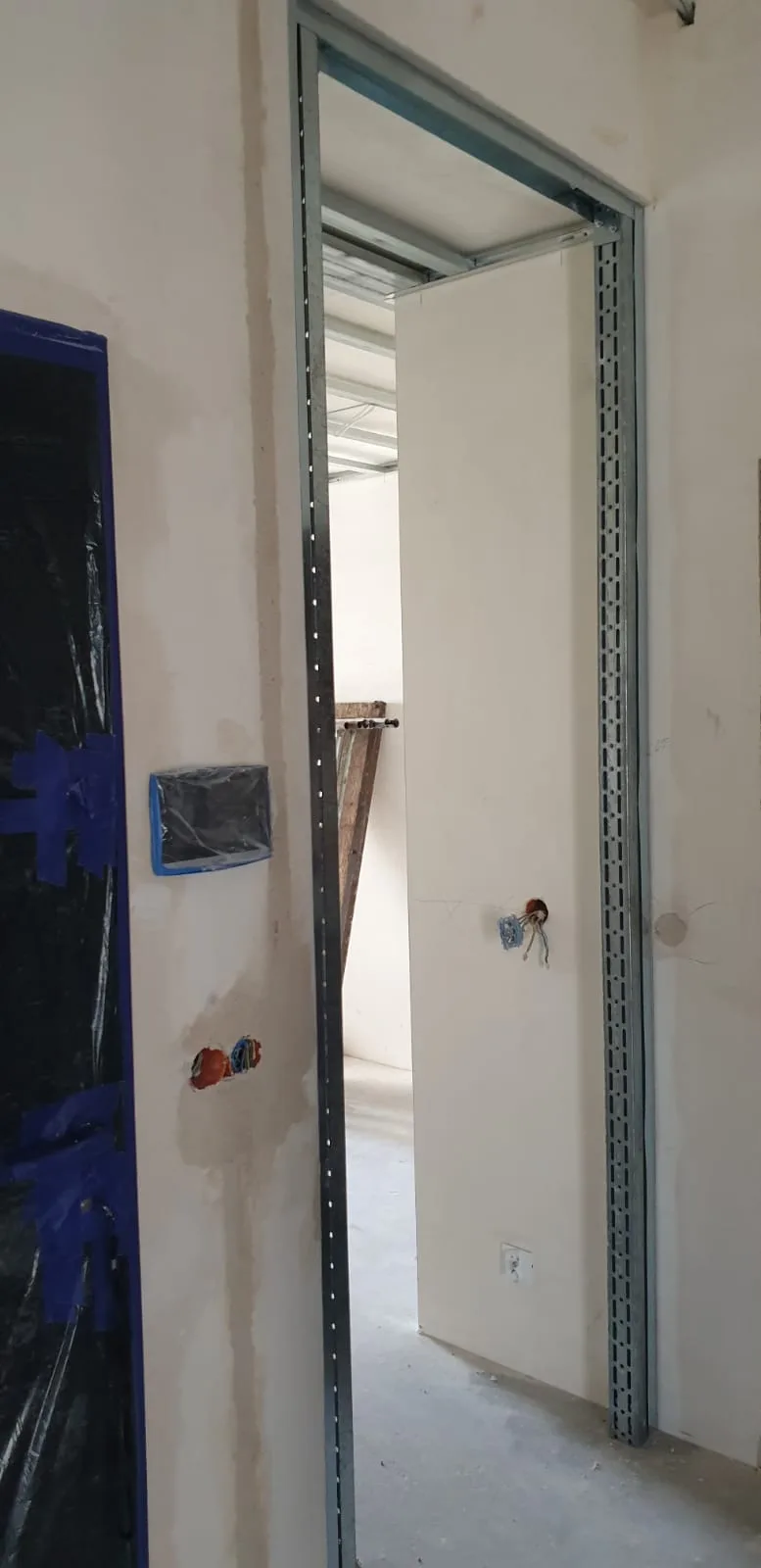 Metal stud C-profile door frame installed during partition wall construction, electrical junction boxes roughed in, blue protective tape on entrance door