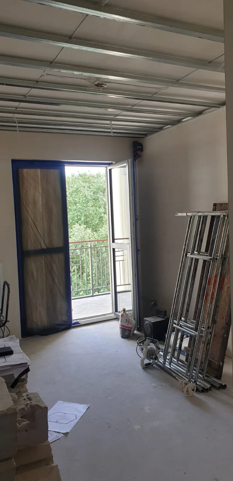 Room during construction with suspended ceiling metal profile grid, French door to balcony with blue protective tape, scaffolding and construction materials stored