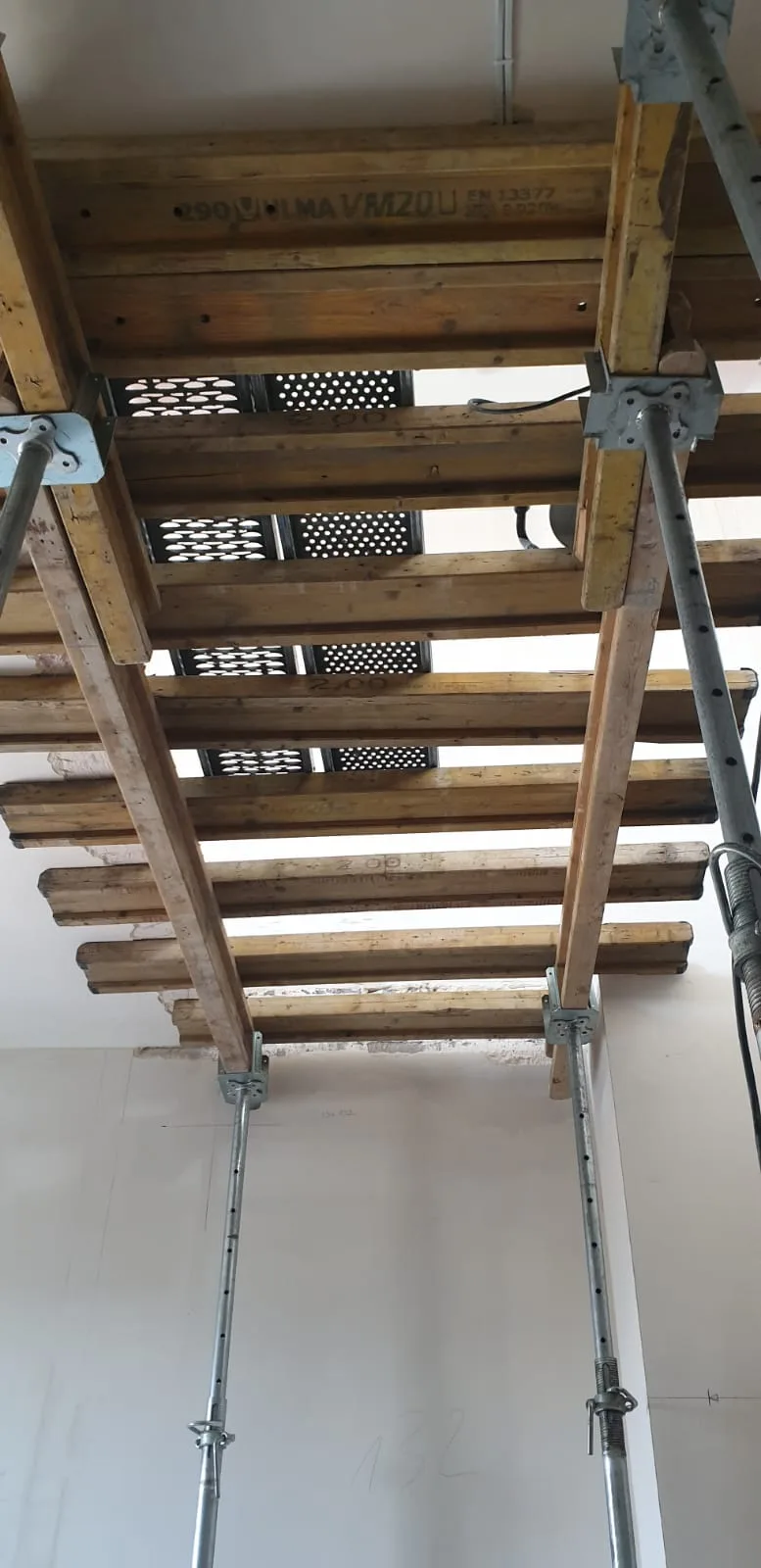 ULMA VM20U formwork system viewed from below showing EN 13377 certified timber beams, perforated metal brackets, and steel acrow props supporting the slab during modification